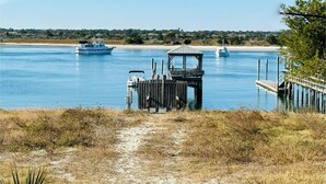 A coastal path leading to a wooden dock with a small pavilion, boats on the water, and a sandy beach in the background.