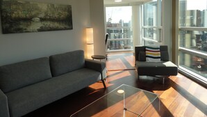 Ein modernes Wohnzimmer mit grauem Sofa, einem Glastisch und Blick auf die Stadt durch große Fenster.