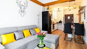 A modern living room with a grey sofa, yellow cushions, a glass coffee table, and a vase with flowers. There is a kitchen in the background with wooden flooring and a dining area with black chairs.