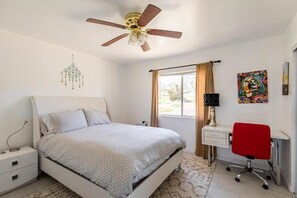 A bedroom with a bed, a desk with a chair, a ceiling fan, a window with curtains, and a wall-mounted artwork. - Relaxing Retreat Close to Park, Hot Tub, Hammocks, Private, Lush Trees (Joshua Tree)
