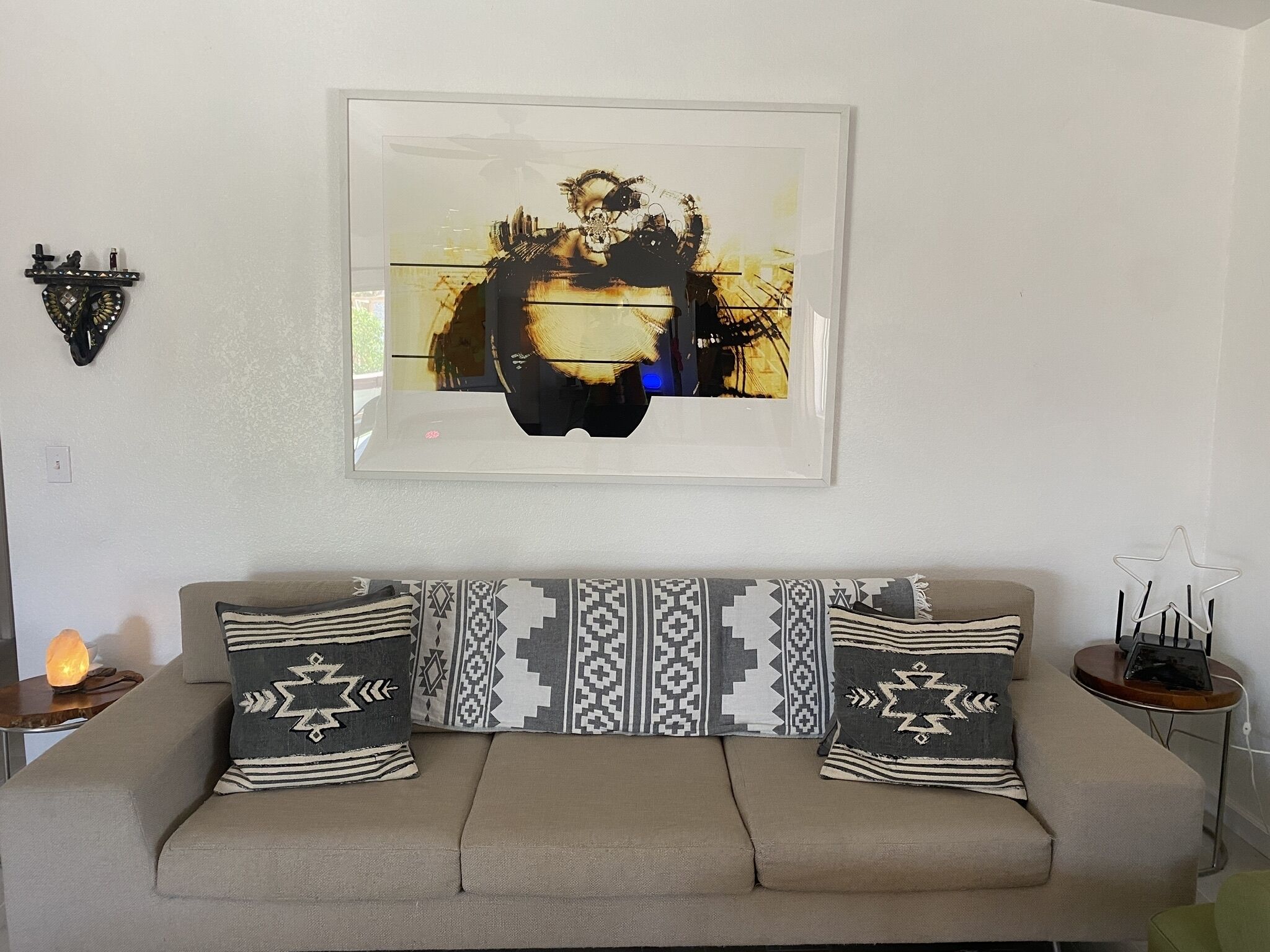 A living room with a beige sofa, two patterned throw pillows, a framed abstract painting, a small table with a lamp, and a wall-mounted star decoration.