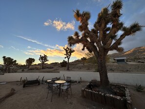 A desert landscape with a wooden deck, metal chairs, and a small table. - Relaxing Retreat Close to Park, Hot Tub, Hammocks, Private, Lush Trees (Joshua Tree)