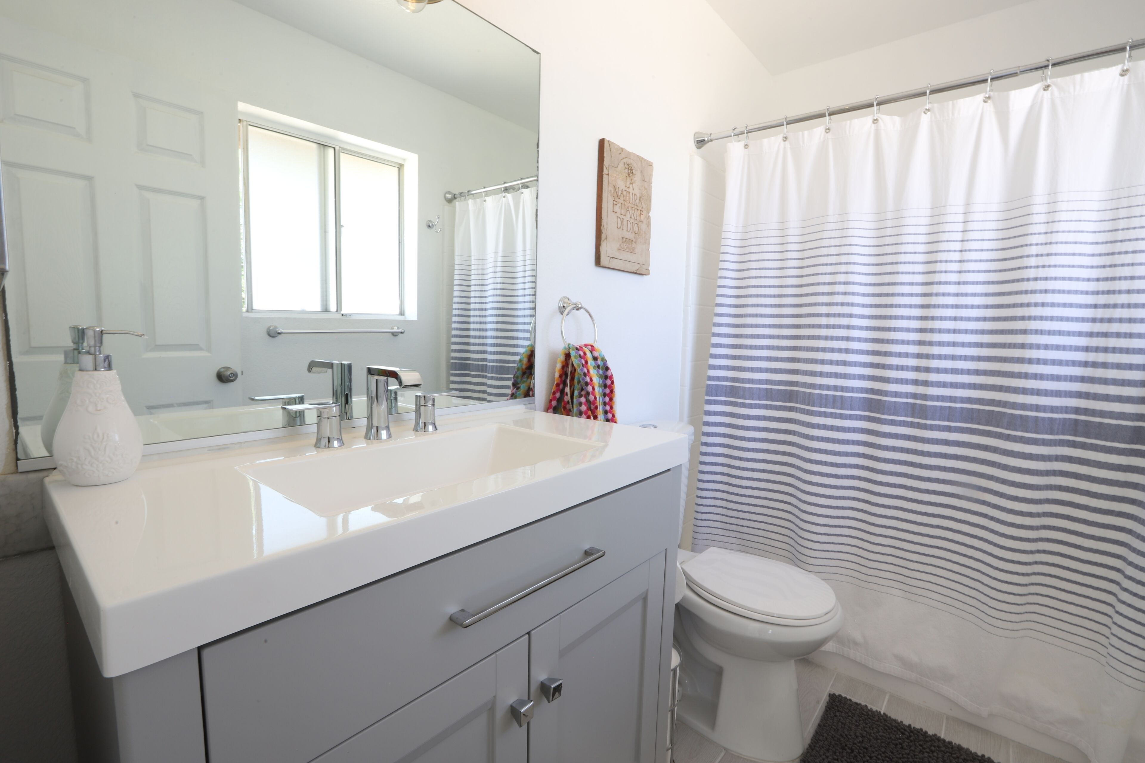 Une salle de bain avec un lavabo blanc, un miroir et des toilettes. On y trouve un rideau de douche à rayures et un petit tapis sur le sol.