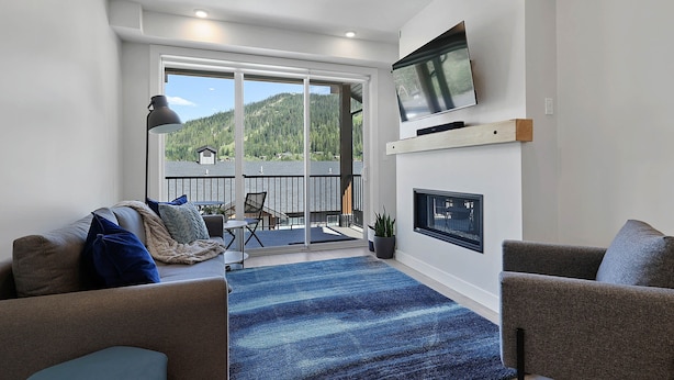 A modern living room with a sofa, a TV mounted on the wall, a fireplace, and a view of a balcony with outdoor furniture. - Townhome with Garage, 3 Bedrooms and Hot Tub (Sun Peaks)