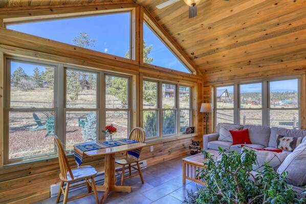 A spacious room with large windows, a wooden ceiling, a dining table with chairs, a sofa, and a small table with a lamp.