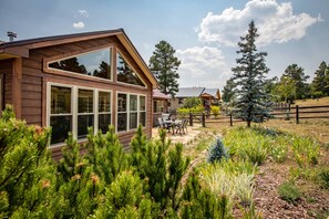 Eine Holzhütte mit großen Fenstern, einer Terrasse mit Gartenmöbeln und einem eingezäunten Garten mit verschiedenen Pflanzen und Bäumen.