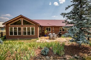 A two-story wooden cabin with a red roof, a patio with outdoor furniture, and a garden with various plants.