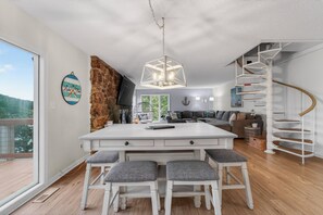 Une cuisine moderne avec un îlot central, des tabourets de bar blancs, un escalier en colimaçon et une grande fenêtre donnant sur un espace verdoyant.