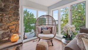 Une pièce avec un mur en pierre, un fauteuil en osier avec des coussins, une table avec une plante et une vue sur les arbres à travers de grandes fenêtres.