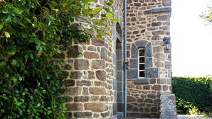 Ein Steinhaus mit einem kleinen Fenster und Stufen, die zu einer Tür führen. Auf den Stufen stehen Blumentöpfe.