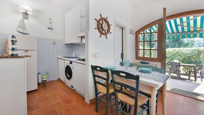 A compact kitchen with a washing machine, a small dining area with a table and chairs, and a view of the garden through a window. - Casa Le Palme with an equipped outdoor area (Porto Azzurro)