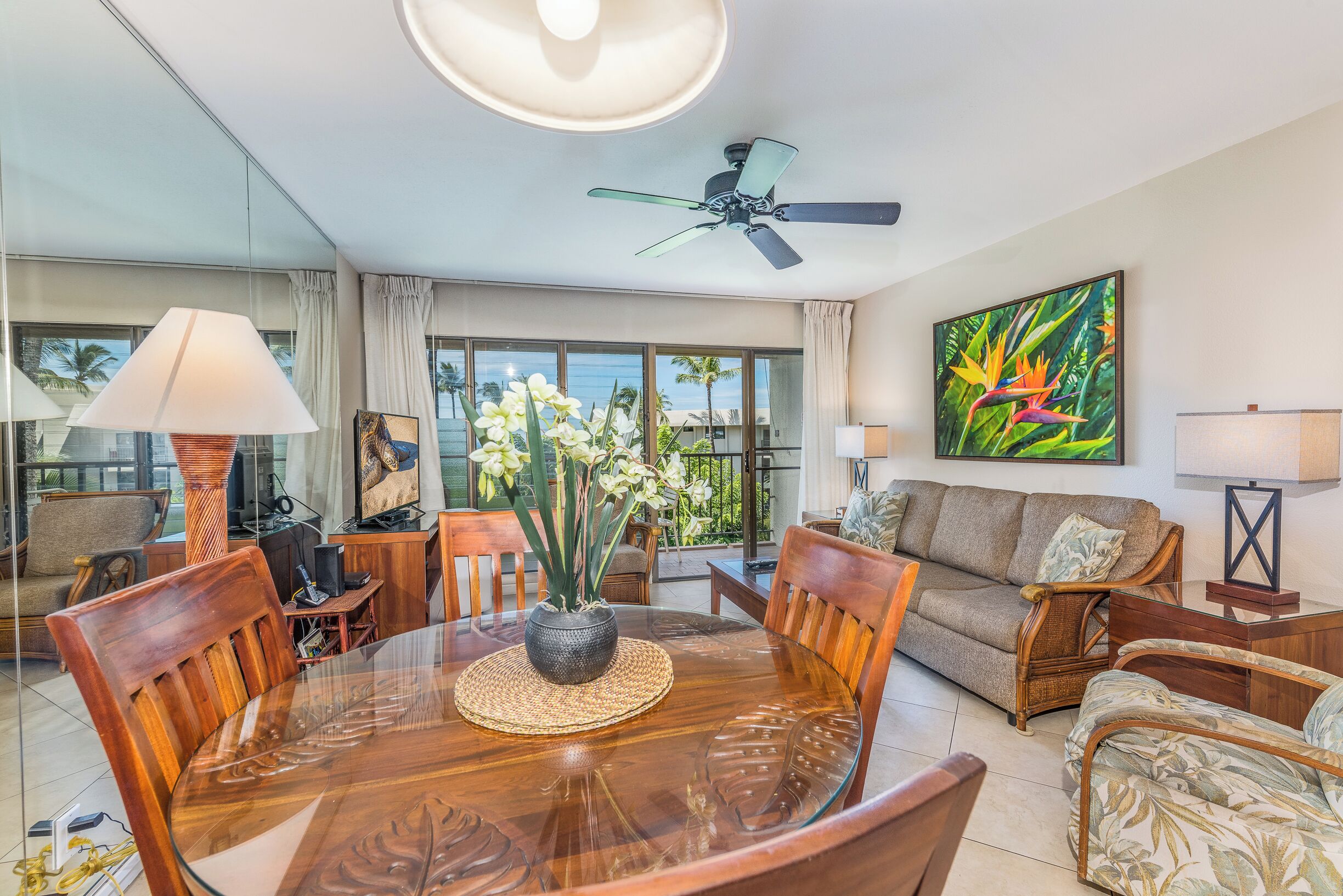A spacious living room with a glass dining table, comfortable seating, a ceiling fan, and a large window offering a view of palm trees.