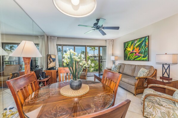 A spacious living room with a glass dining table, comfortable seating, a ceiling fan, and a large window offering a view of palm trees.
