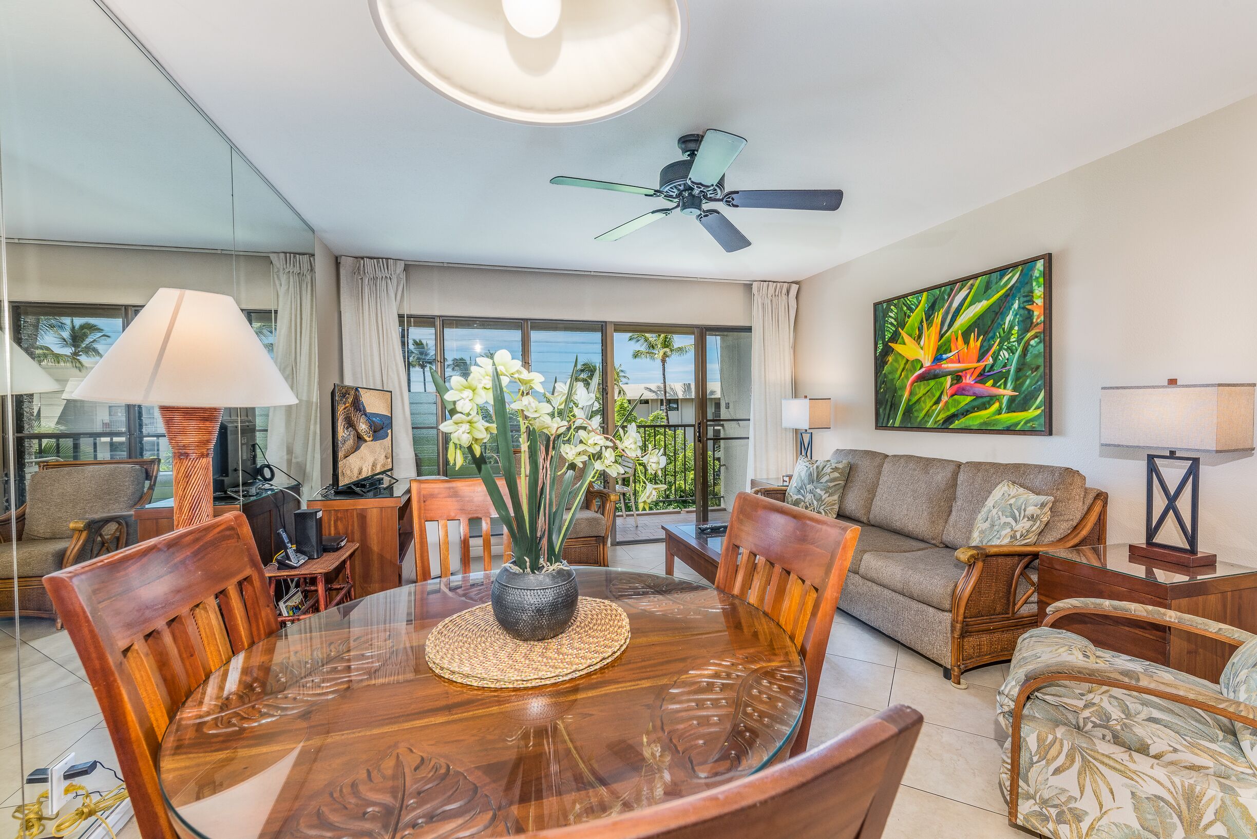 A spacious living room with a glass dining table, comfortable seating, a ceiling fan, and a large window offering a view of palm trees.