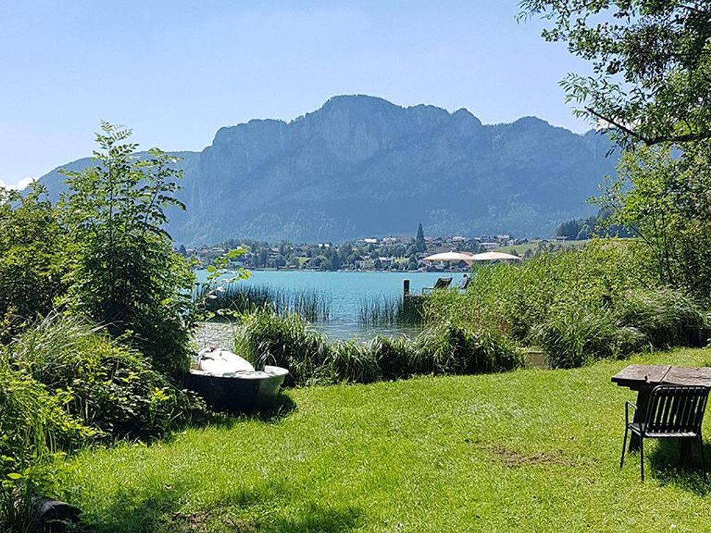 Vista para um lago com montanhas ao fundo, uma área gramada e um banco.
