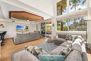 A spacious living room with a large gray sectional sofa, a flat-screen TV mounted on the wall, and a large window offering a view of palm trees and a house.