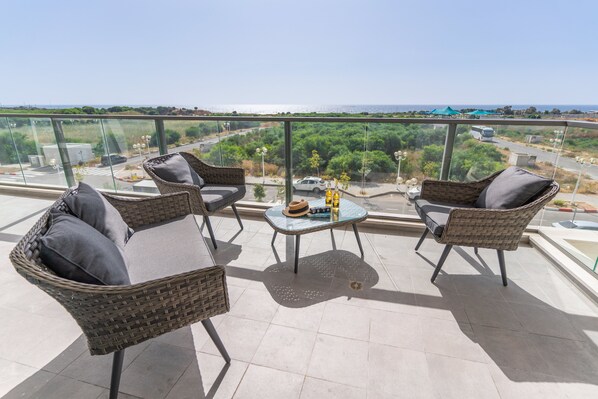 A balcony with wicker furniture, a small table, and a view of the sea and greenery.