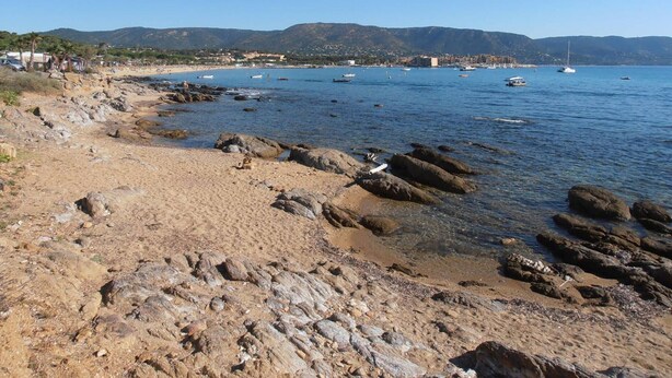 Uma praia rochosa com águas azul-claras, barcos e uma paisagem urbana ao longe.