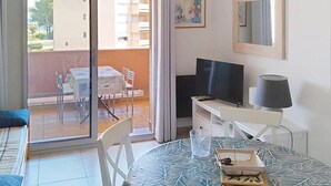 Quarto com porta de vidro deslizante que dá para uma varanda externa com mesa e cadeiras. Há uma cama com travesseiro azul, uma televisão, um abajur e uma pequena mesa com um tabuleiro.