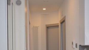 Ein schmaler Flur mit Heizkörper, einer Tür am Ende und einem weißen Kleiderschrank mit Lüftungsschlitzen.