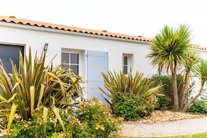 Un edificio blanco con techo de tejas, una puerta azul y un jardín con diversas plantas y una palmera.