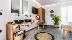 Un baño moderno con dos lavamanos grandes, un tocador de madera, un espejo, un televisor y una planta en maceta.
