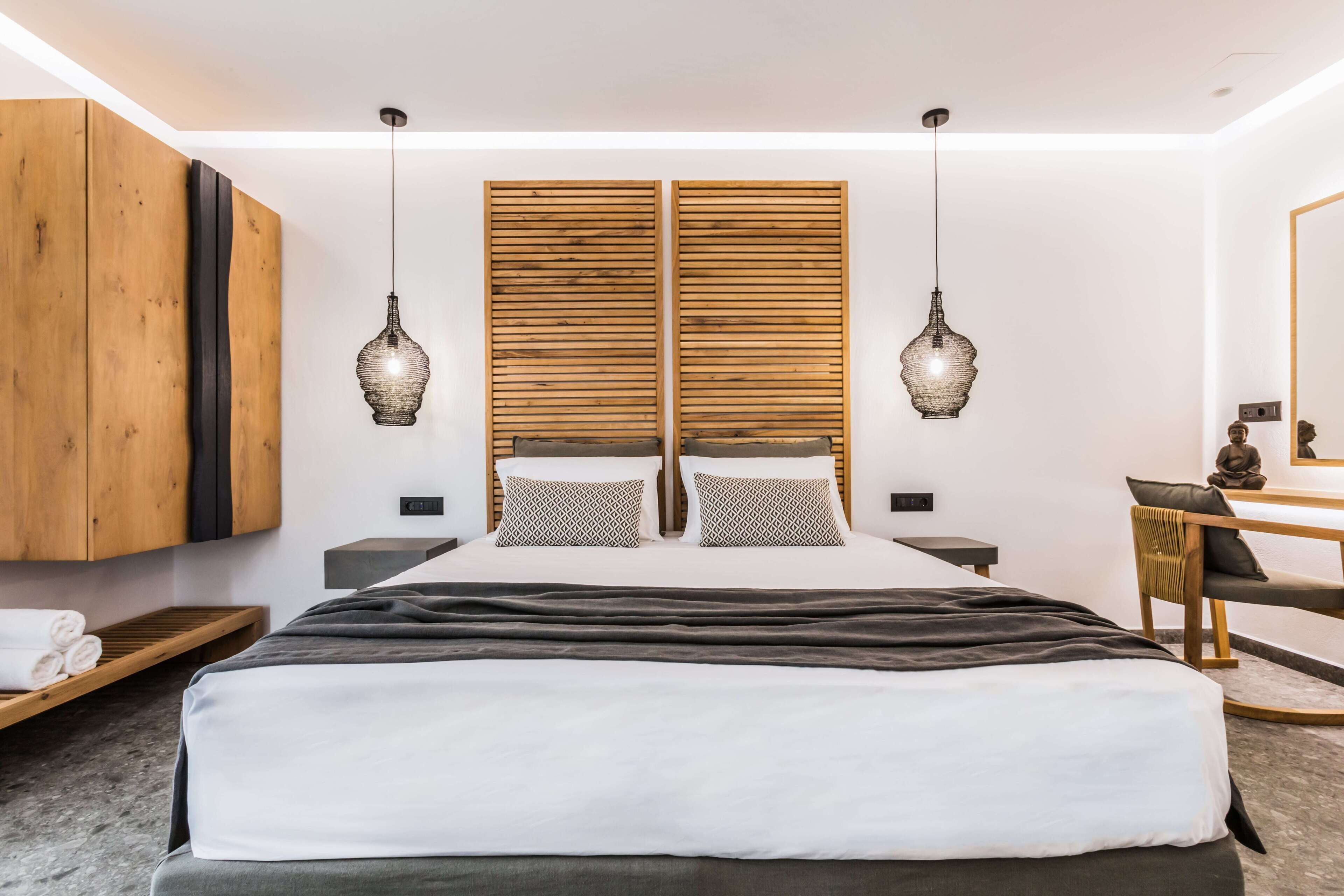 A modern bedroom with a large bed, wooden headboard, two pendant lights, a wooden wardrobe, a desk, and a chair.