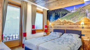 A bedroom with a wooden headboard, a bed with patterned bedding, a window with curtains, and a mural of a mountainous landscape on the wall. - Appartamento Livigno (Livigno)