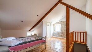 Un dormitorio con techo inclinado, vigas de madera, cama con colcha estampada, pared de piedra y escalera de madera.