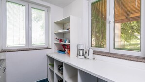 Una cocina con armarios blancos, una encimera y dos ventanas con persianas. Se ven estantes con utensilios de cocina y una cafetera sobre la encimera.