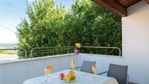 Un balcón con una mesa dispuesta para dos personas, cubierta con un mantel blanco, y dos sillas. Hay tres vasos de una bebida amarilla con rodajas de naranja sobre la mesa. Una maceta con flores amarillas está en la barandilla.