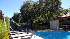 Una piscina con sillas de descanso y sombrillas, rodeada de árboles y un muro de piedra.