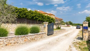 Un camino rural que conduce a una propiedad con muros de piedra, una reja, un coche estacionado y un cartel que indica un precio de 62 euros por noche.