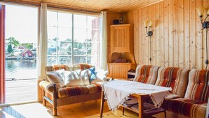 A cozy cabin with a wooden interior, a sofa, a table, and a view of a water body and houses outside.
