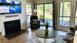 Una sala de estar con chimenea, televisor montado en la pared, un sofá cómodo, una mesa de centro y vistas al exterior a través de amplios ventanales.