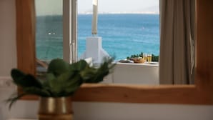 A view through a window with a potted plant on a windowsill, overlooking a body of water and a distant shoreline. - Avali Deluxe Suite II (Naxos)