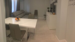 Una sala de estar moderna con un sofá blanco, una mesa de comedor con sillas, un pequeño armario blanco e iluminación en el techo.