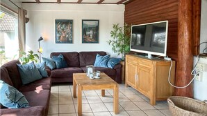 En stue med en brun sofa, et træbord og et fjernsyn på et træskab. Der er to billeder i rammer på væggen og en plante.