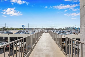 Un puente peatonal que conduce a un área de estacionamiento con varios coches, cielo despejado y edificios al fondo.