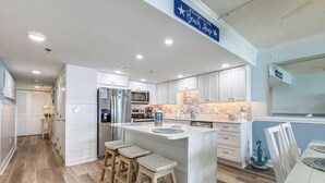 A modern kitchen with white cabinetry, a large island with stools, a microwave, and a refrigerator. - This absolutely beautiful 3 bedroom, 2 bath condo completely remodeled into a stunning beach home. (Ocean City)