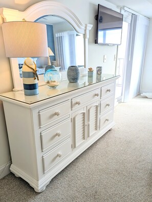 Une commode blanche avec des tiroirs et un miroir, une lampe, des vases décoratifs et un téléviseur fixé au mur.