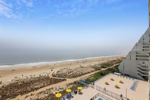 Un hotel de gran altura con piscina en la azotea, vistas a la playa y una zona de playa de arena con sombrillas y tumbonas.