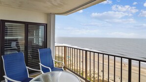 Un balcón con una mesa de vidrio y sillas azules, con vista a una playa con barandilla.