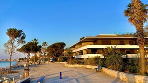 A beachside promenade with a wooden deck, palm trees, and a multi-story building with balconies. - Haven of peace with infinity pool overlooking the sea (Villeneuve-Loubet)
