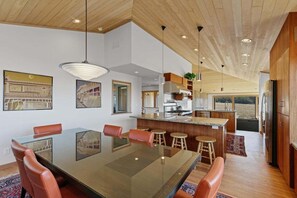 A modern kitchen with a central island, wooden cabinets, and a dining area with a glass table and leather chairs. - Kyuka | Asian-Inspired Coastal Retreat (Sea Ranch)