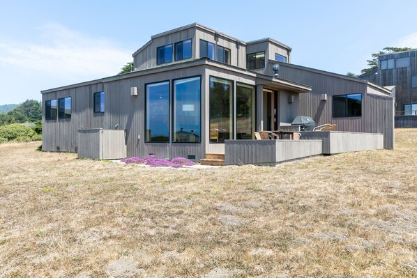 Una casa moderna de dos pisos con ventanas amplias, un patio con mobiliario exterior y vistas del paisaje circundante.