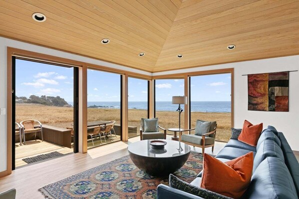 A modern living room with a large window, a round coffee table, and a sofa with cushions. - Kyuka | Asian-Inspired Coastal Retreat (Sea Ranch)