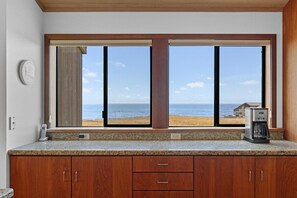 A kitchen with a large window overlooking a beach, a coffee maker on the counter, and wooden cabinets. - Kyuka | Asian-Inspired Coastal Retreat (Sea Ranch)