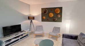 Un salon équipé d’un téléviseur à écran plat, de deux fauteuils gris, d’une petite table ronde et d’un lampadaire.