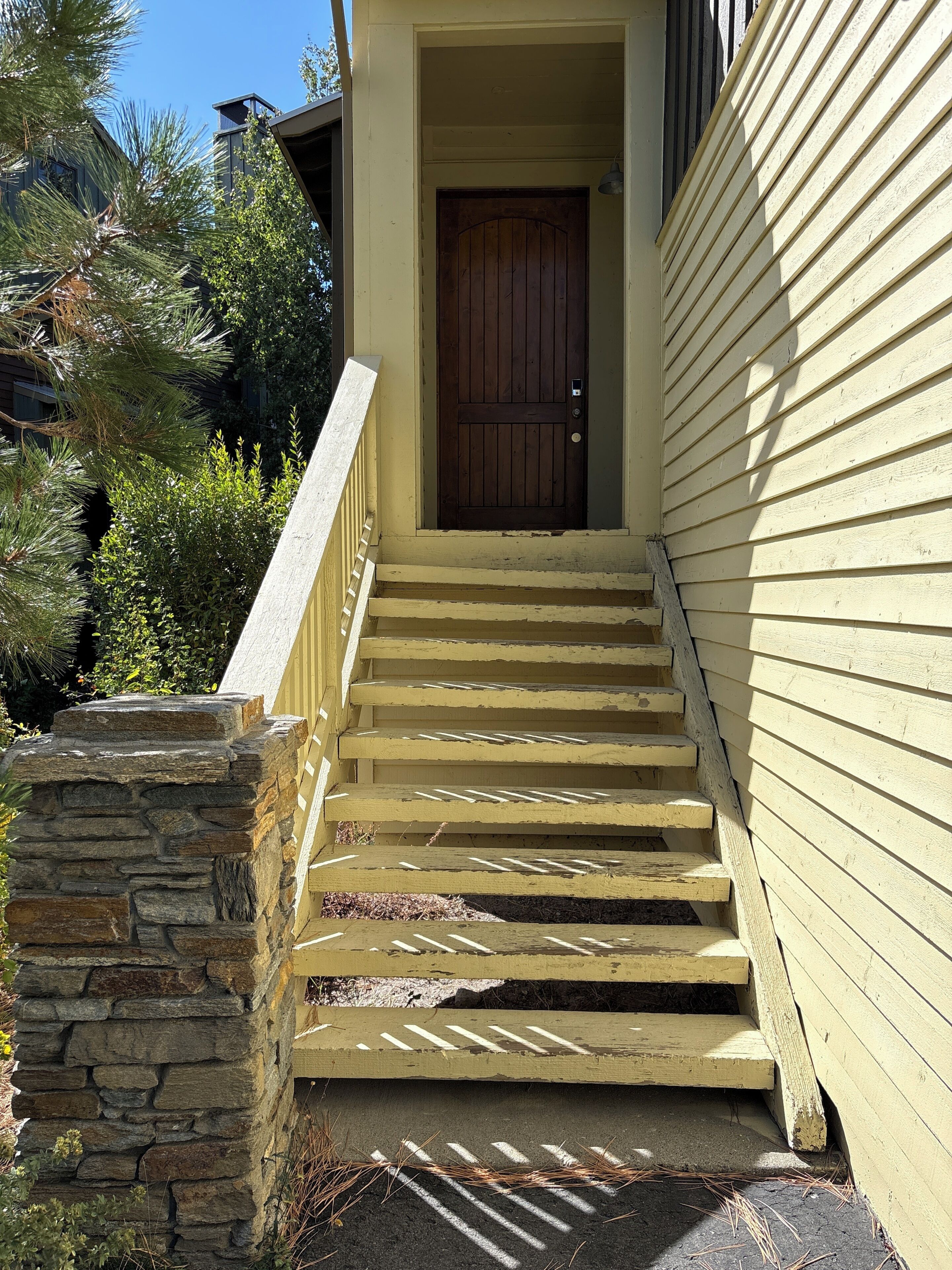 Un escalier en bois mène à une porte en bois, avec un mur de pierre d’un côté et une maison recouverte de bardage jaune de l’autre.